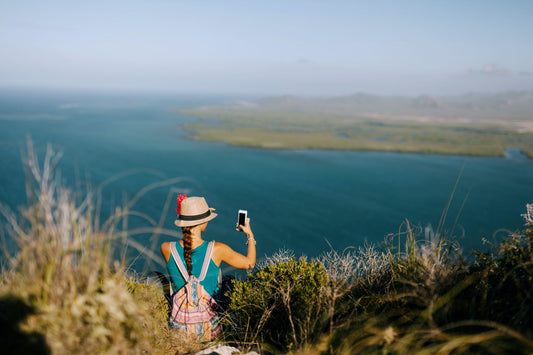 Female tourist with backpack and hat taking a picture of the sea and using her eSIM to share with firends
