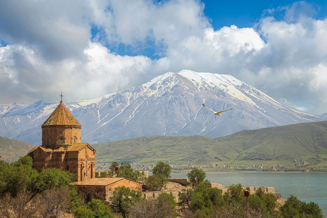 Best eSIM for Armenia; photo of a  scenic landscape in Armenia featuring a snow-capped mountain range, a blue lake, and a historic stone church in the foreground.