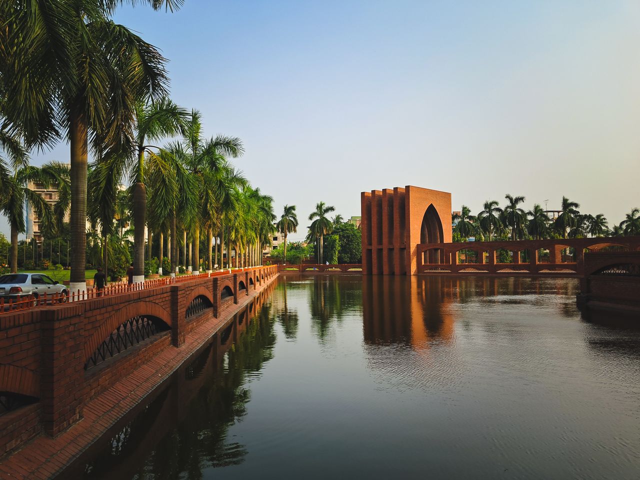 Best eSIM for Bangladesh; a scenic view of a large reflective water canal in Bangladesh with a tall arched brick monument at the center, lined with tall palm trees and a brick walkway.