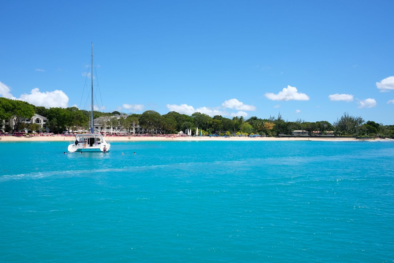 Best Bardados eSIM for travel; a coastal view in Barbados with bright turquoise water in the foreground, a white sailboat near the center-left, a sandy beach lined with green trees and houses.