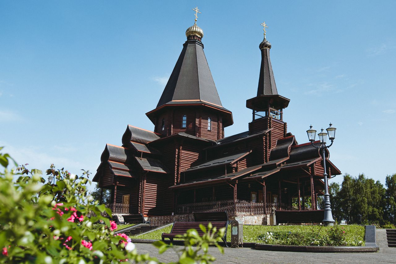Best eSIM for Belarus; photo of a large dark brown wooden church in Belarus with multiple steep dark roofs and golden onion domes, with greenery and flowers in the foreground.