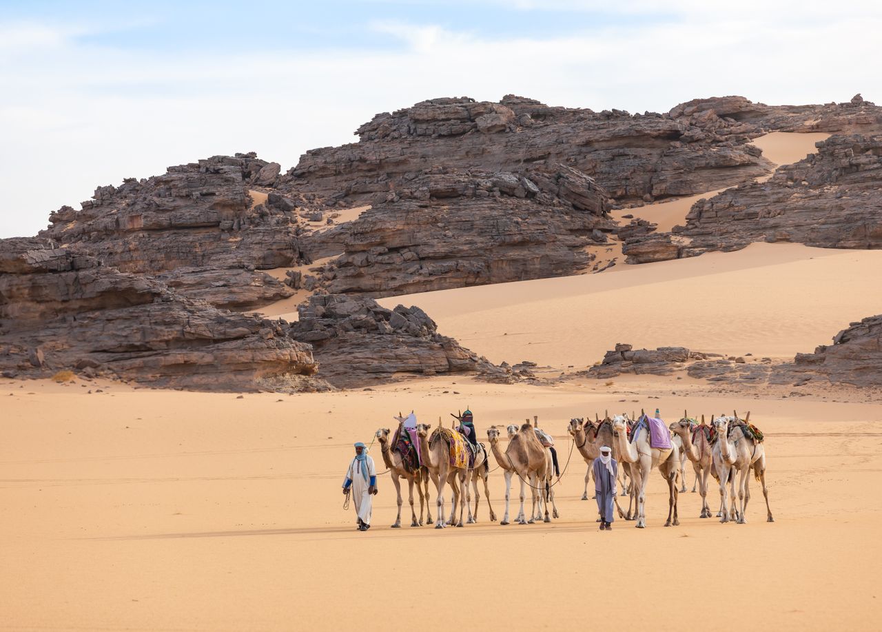 Best Chad eSIM for travel; a desert landscape in Chad with sand dunes, rocky cliffs, and a caravan of camels being led by two people under a pale blue sky.