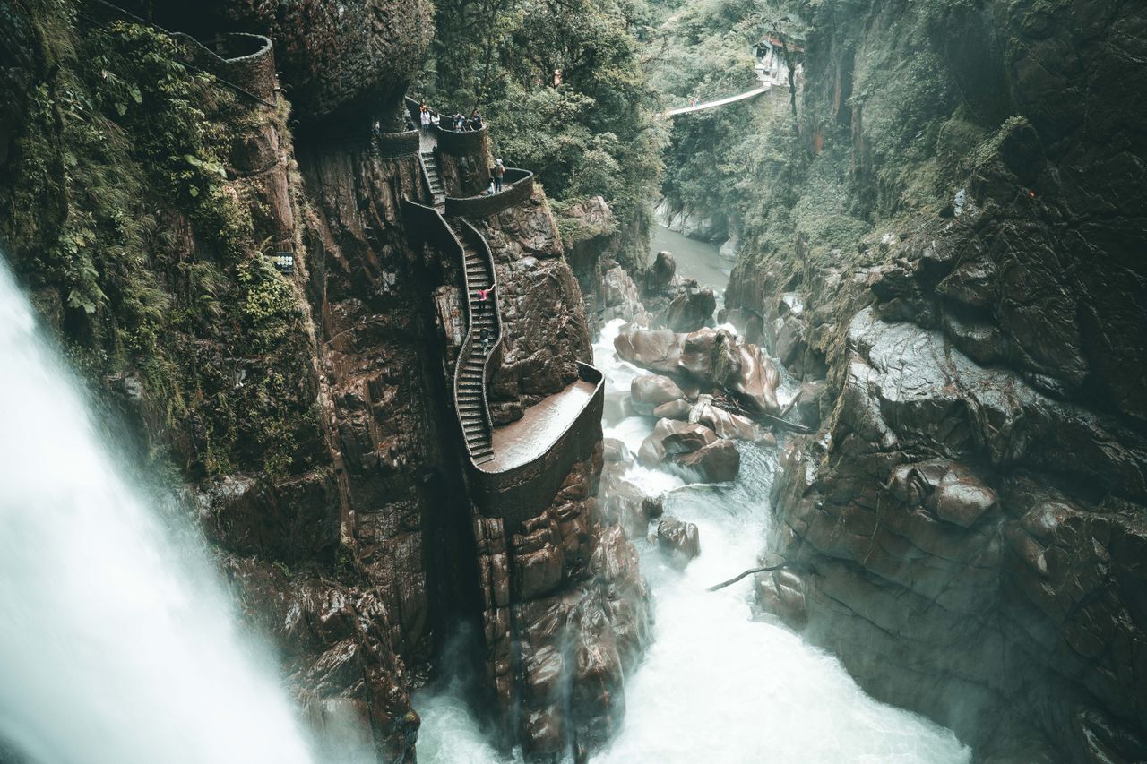 Best Ecuador eSIM for travel; a dramatic natural canyon scene with a waterfall on the left, rocky cliffs, a rushing river, and a narrow elevated walkway or bridge winding through the gorge in Ecuador.