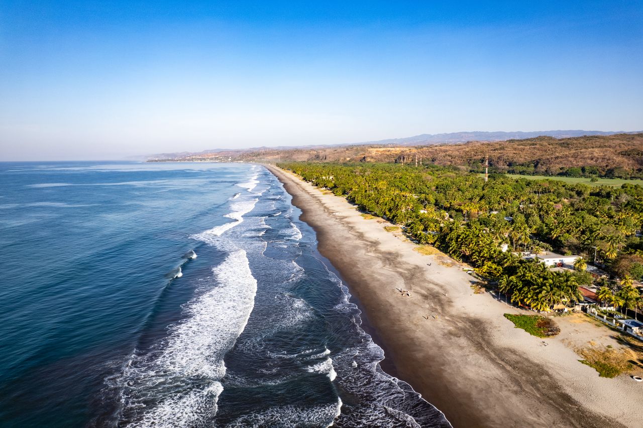 Best El Salvador eSIM; an aerial view of a long sandy beach running parallel to a deep blue ocean in El Salvador, with white waves breaking along the shore and dense green palm trees.