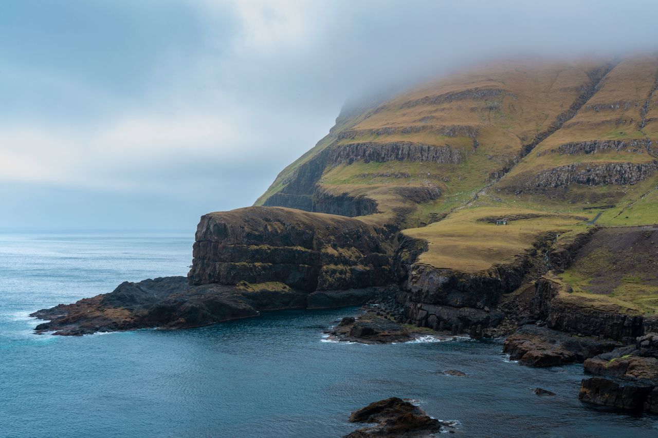 Best Faroe Islands eSIM; a dramatic coastal landscape with steep grassy cliffs rising from dark rocky shores into a misty blue sky over calm water.