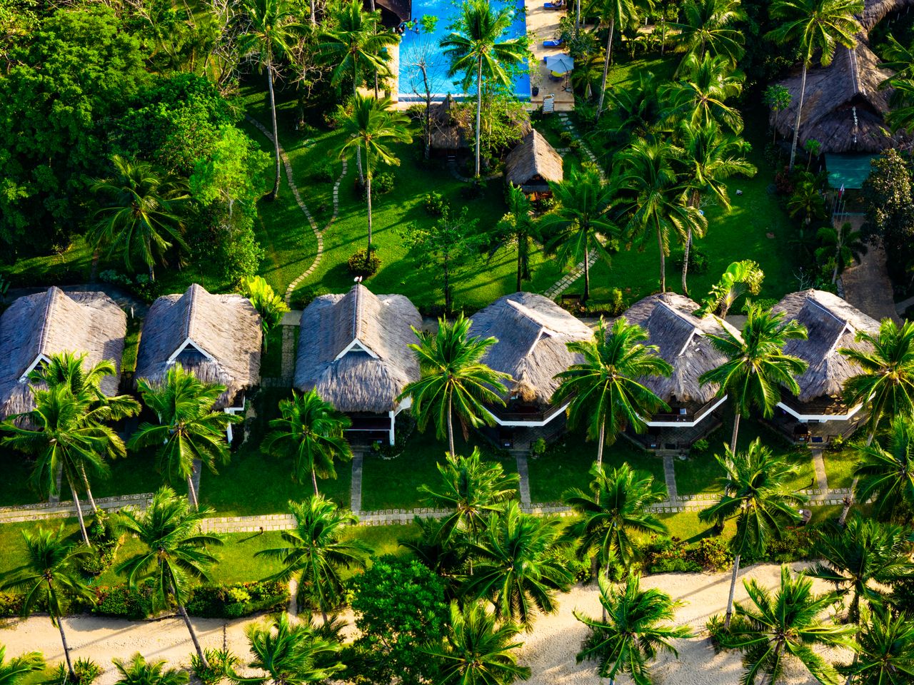 Best eSIM for Fiji; aerial view of a tropical resort in Fiji with thatched-roof bungalows, palm trees, green lawn, and a blue swimming pool.