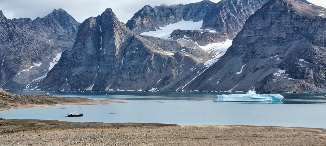Best eSIM for Greenland travel; a dramatic mountain landscape with steep rocky peaks, patches of snow and ice, a calm blue bay in Greenland.