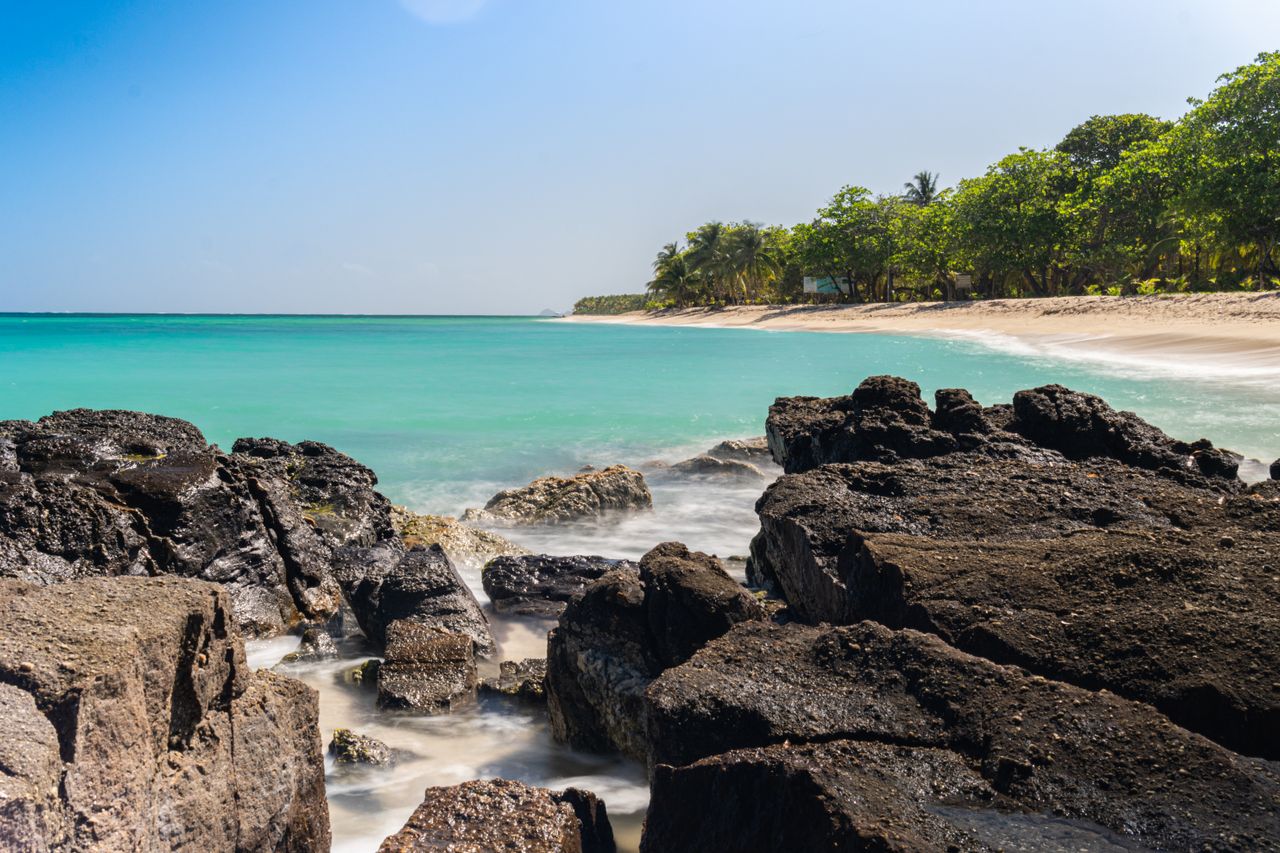 Best Grenada eSIM for travel; a tropical beach in Grenada with turquoise ocean water, dark volcanic rocks in the foreground, a sandy shoreline, and green trees.