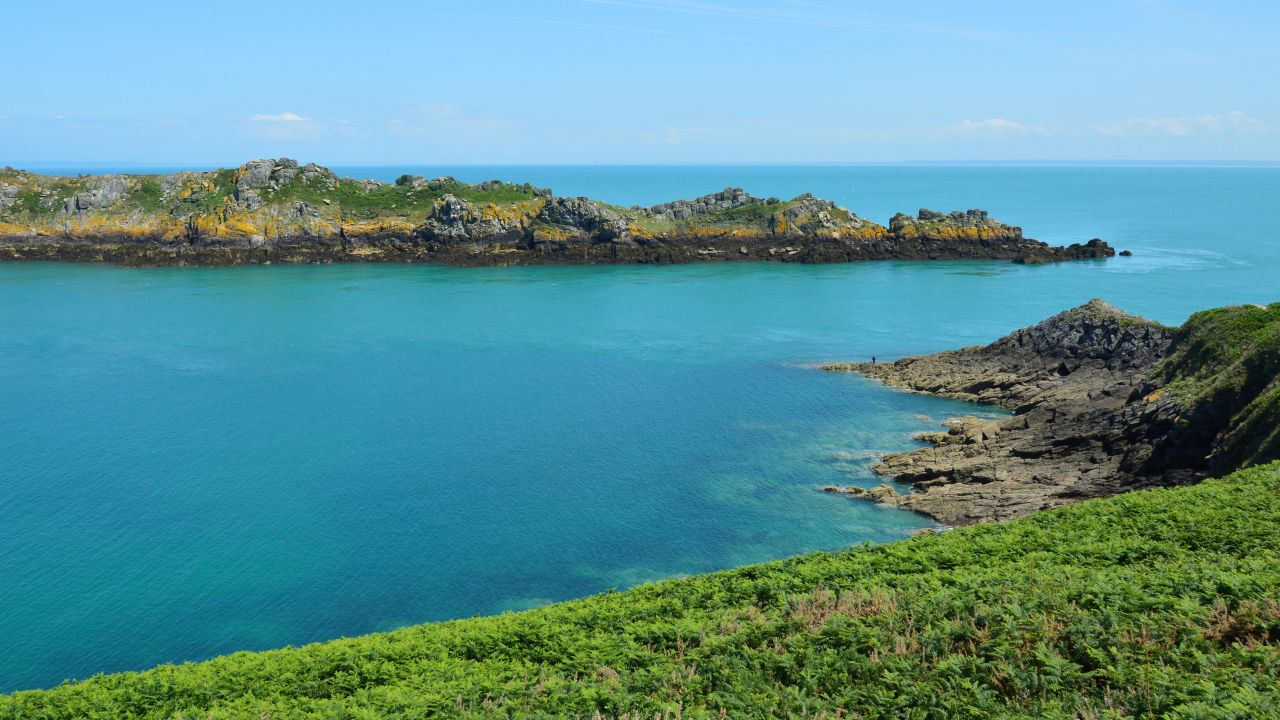 Best eSIM for Guernsey; a scenic coastal landscape in Guernsey with bright turquoise water, rocky shoreline, green grassy foreground, and blue sky.