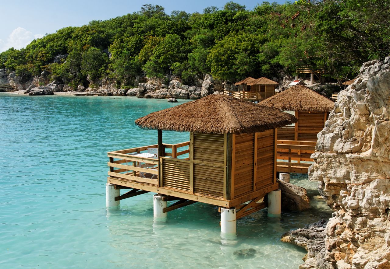 Best Haiti eSIM for travel; overwater wooden cabanas with thatched roofs sit on white pilings in clear turquoise water beside rocky shorelines and a lush green forest.