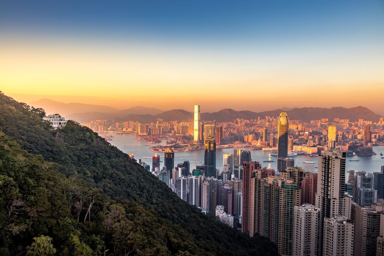 Best eSIM for Hong Kong; scenic view of the Hong Kong skyline at sunset with high-rise buildings, a harbor, mountains in the distance, and a forested hillside in the foreground.