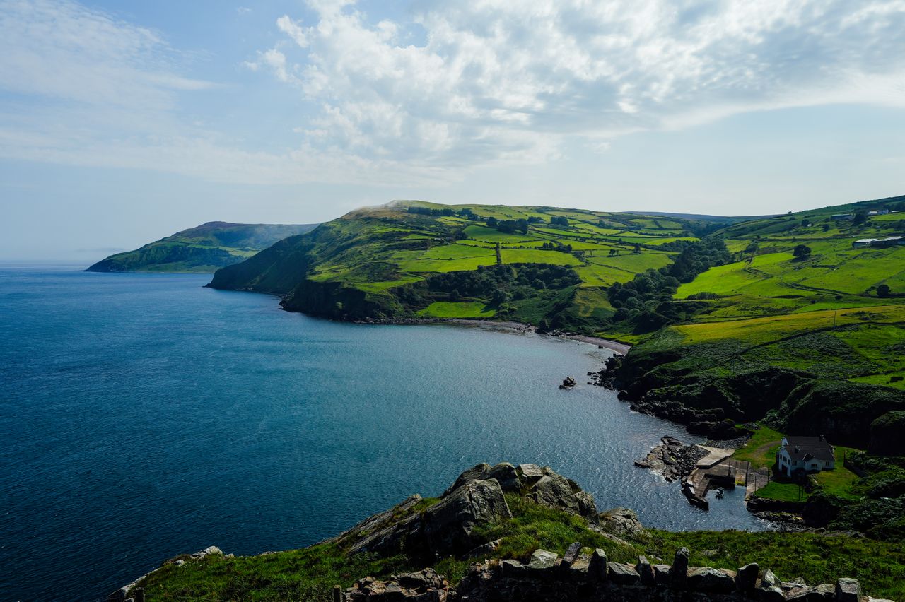Best Isle of Man eSIM for travel; a scenic coastal landscape with deep blue water, green rolling hills, rocky cliffs, and a partly cloudy sky.