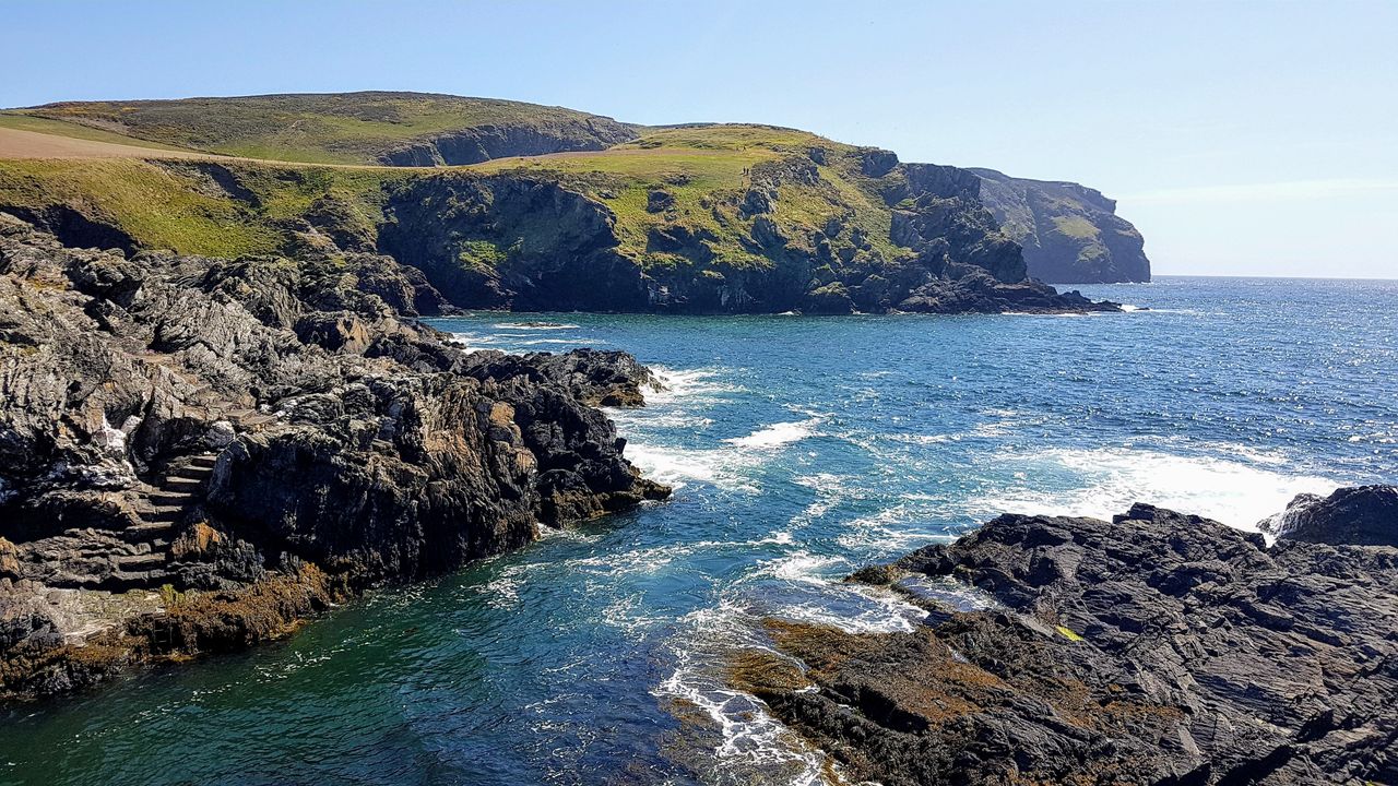 Best eSIM for Isle of Man; a scenic coastal landscape with dark rocky cliffs, green grassy slopes, and blue ocean water under a clear sky.