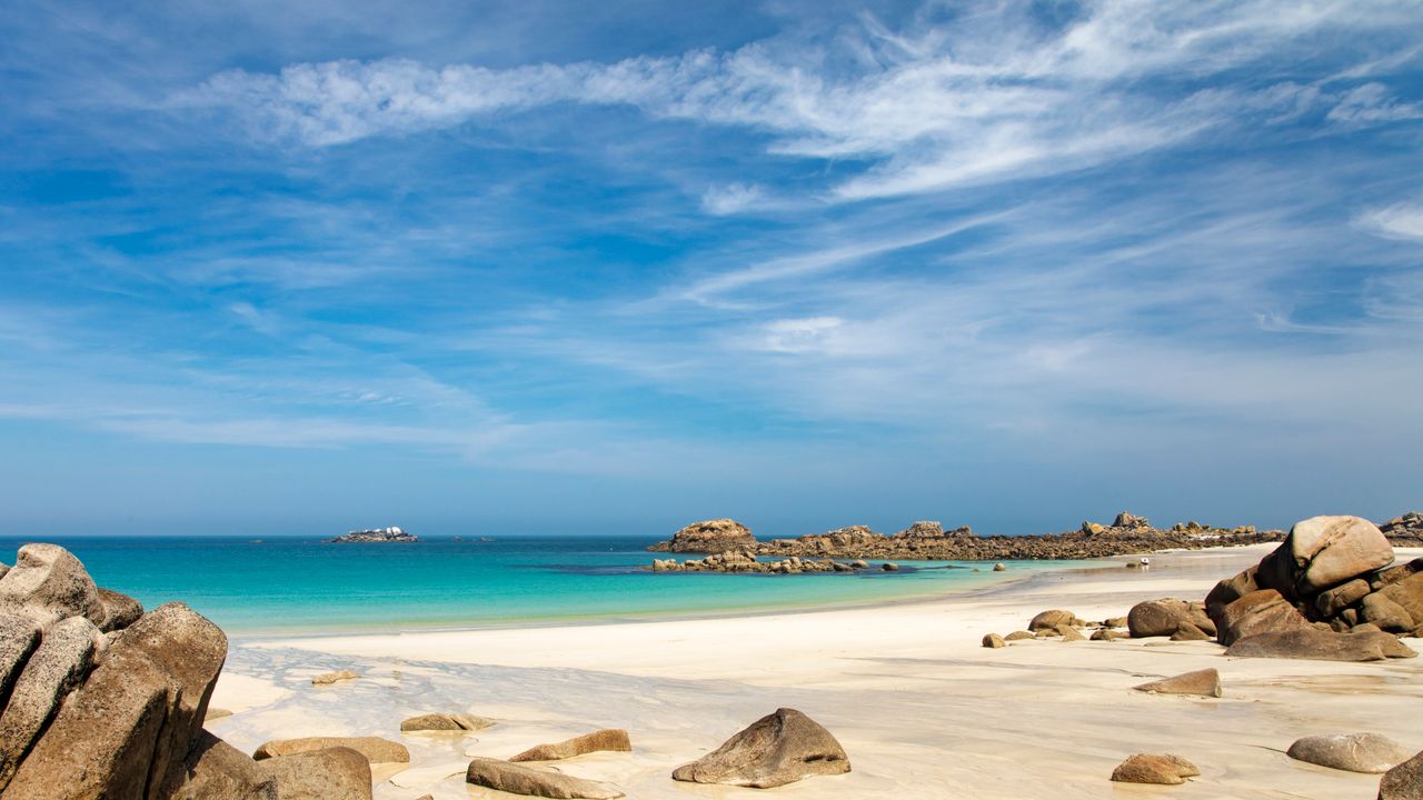 Best eSIM for Jersey; a scenic beach landscape in Jersey with white sand, large brown rocks, turquoise water, and a bright blue sky with wispy clouds.