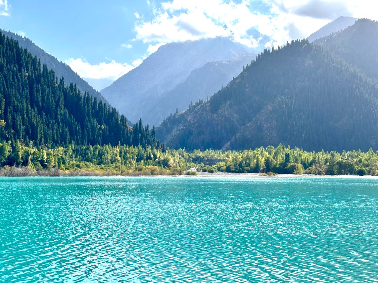 Best Kazakhstan eSIM for travel; a scenic bright turquoise lake in Kazakhstan, bordered by lush green forests and tall mountains under a blue sky with white clouds.
