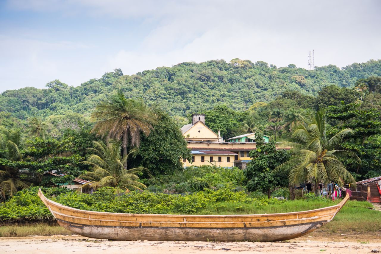 Best eSIM for Liberia; a traditional wooden boat in Liberia resting on a sandy shore in front of dense tropical greenery, palm trees, and a forested hillside.