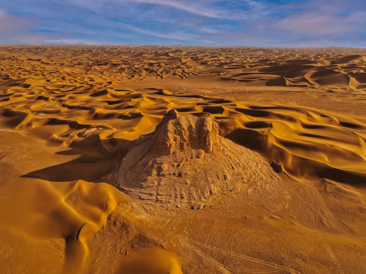 Best Libya eSIM for travel; aerial view of a vast desert landscape in Libya with rolling golden sand dunes and a central rocky outcrop under a blue sky with light clouds.