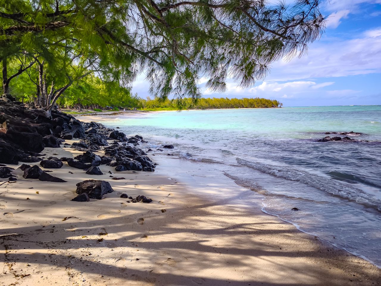 Best Mauritius eSIM for travel; a tropical beach in Mauritius with pale sand, dark volcanic rocks, turquoise water, a line of green trees in the distance, and blue sky with white clouds.