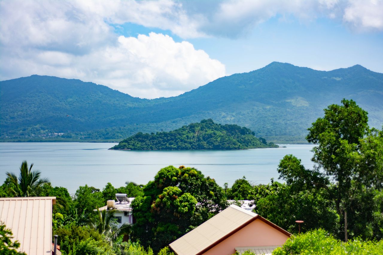 Best Mayotte eSIM; a scenic landscape showing a calm bay in Mayotte with a small green island in the distance, surrounded by blue mountains and lush green trees.