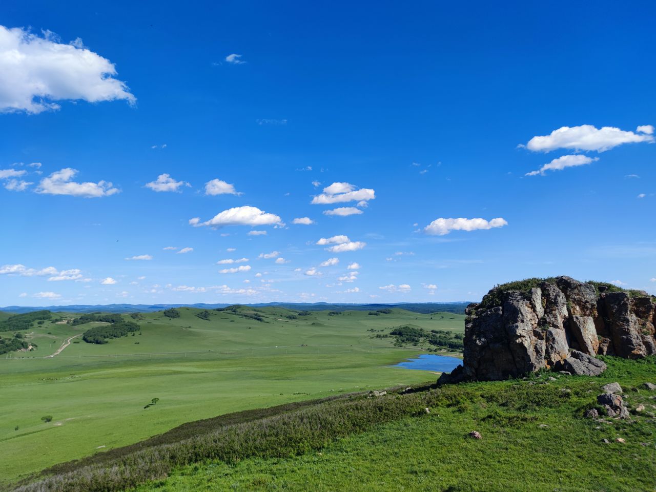 Best Mongolia eSIM: a scenic vast green valley in Mongolia under a bright blue sky with scattered white clouds, featuring a prominent rocky cliff on the right and a small blue lake in the distance.