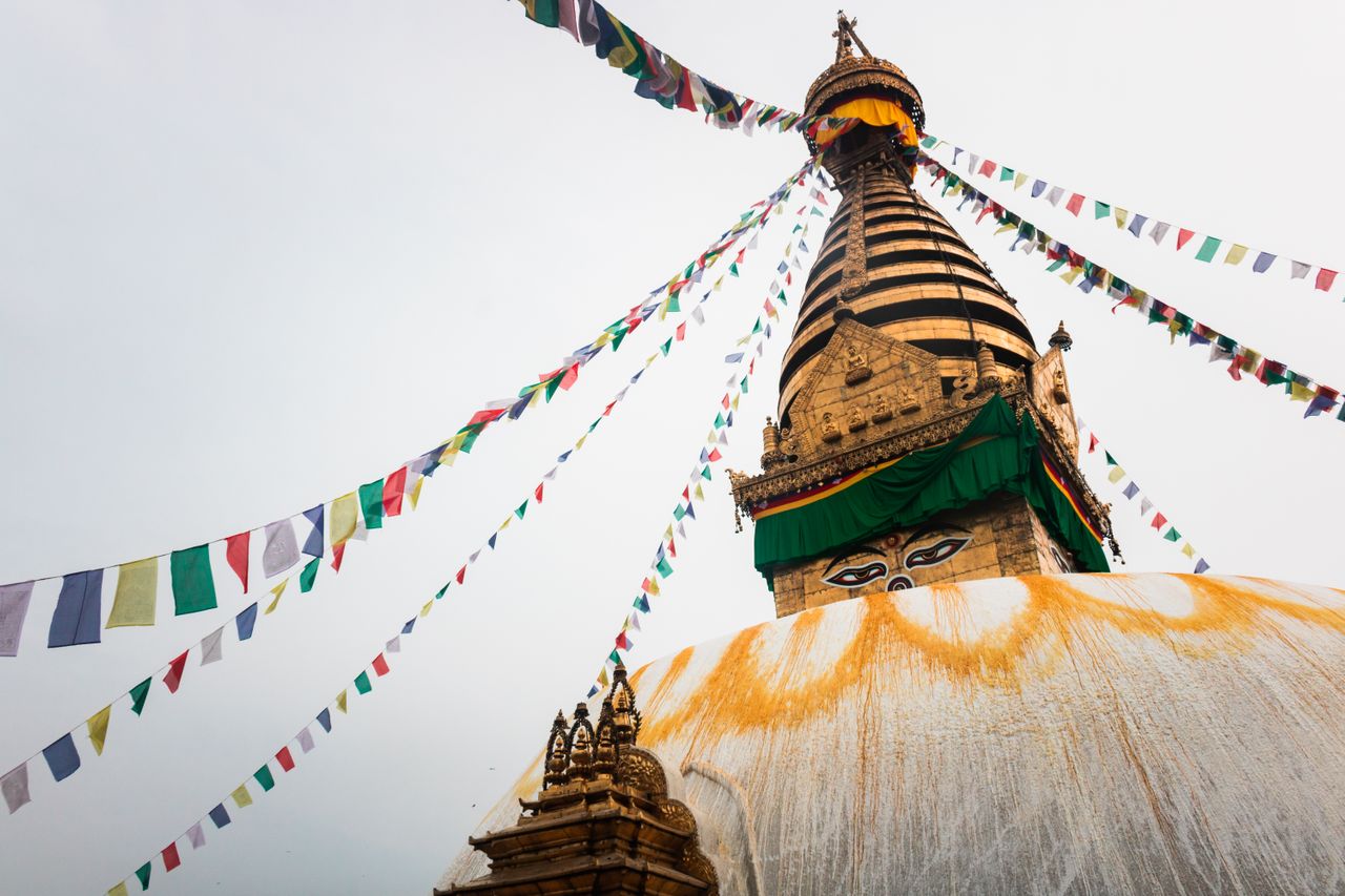 Best eSIM for Nepal; a Buddhist stupa in Nepal with a large white dome marked with orange prayer marks, a striped golden spire, and colorful prayer flags strung across the sky.