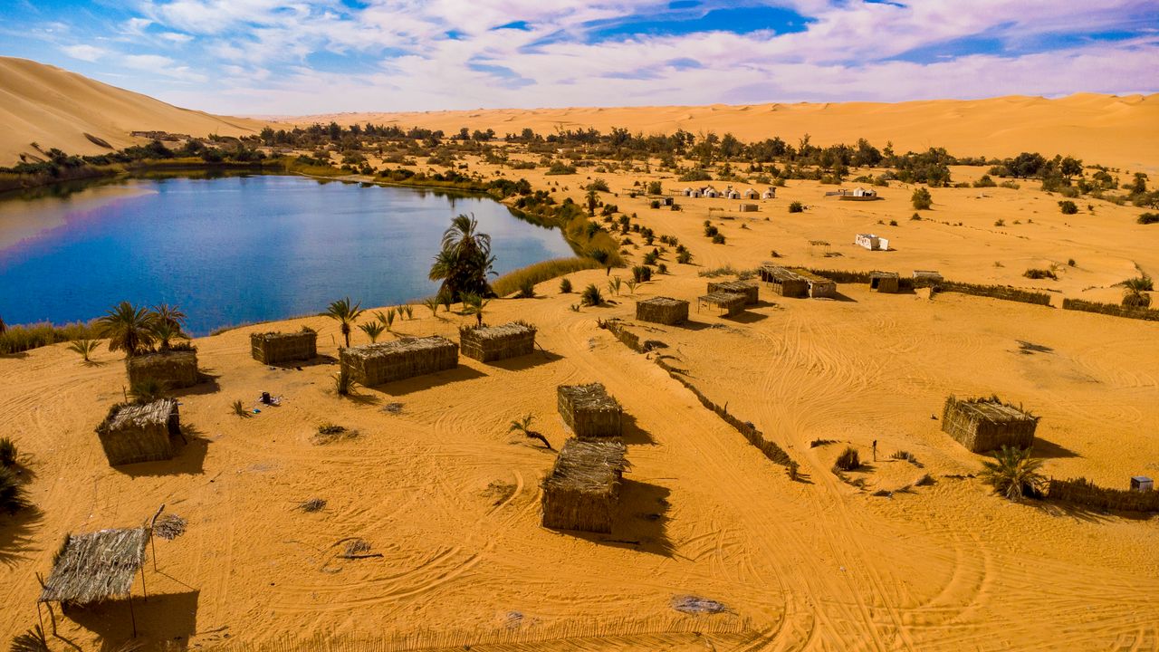 Best eSIM for Niger; aerial view of a desert landscape in Niger with golden sand dunes, a blue lake, scattered palm trees, small rustic buildings.