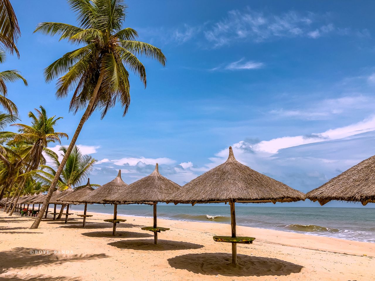 Best Nigeria eSIM for travel: a tropical beach in Nigeria with thatched beach umbrellas on sandy shore, palm trees, blue ocean.