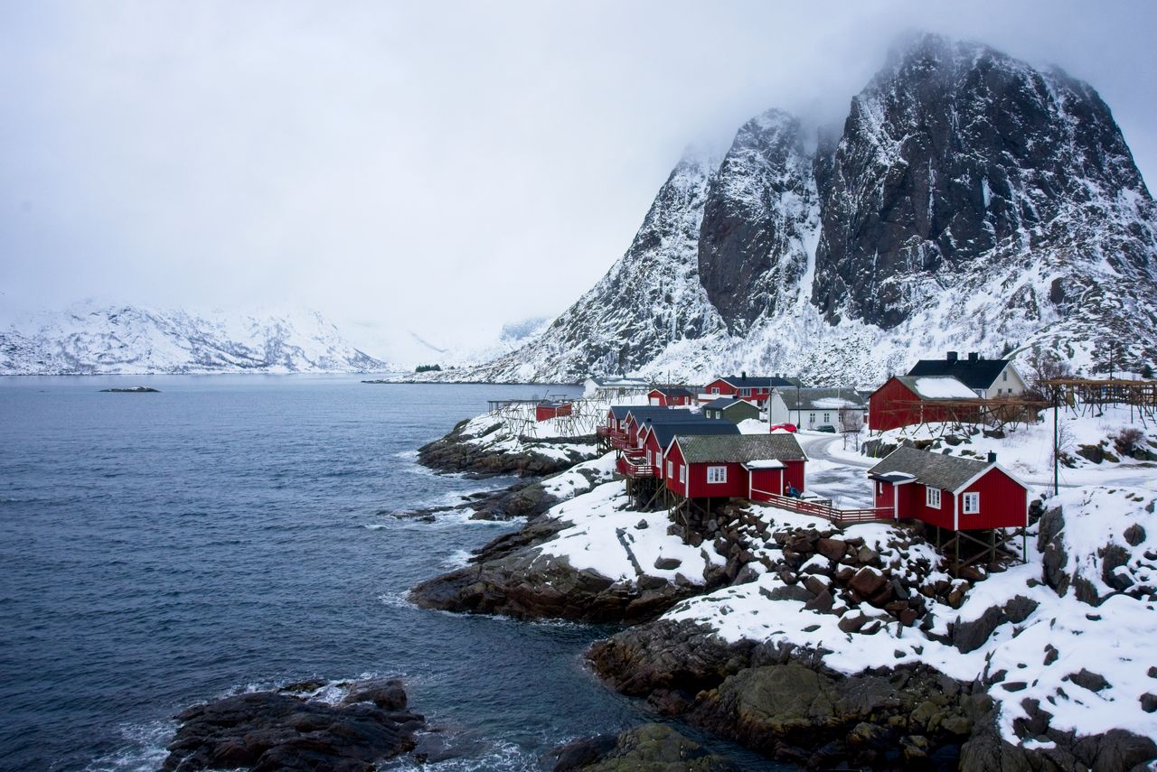 Best Norway eSIM for travel - photo of a snowy coastal landscape with small red cabins beside a rocky shoreline, set against a large dark mountain in Norway.