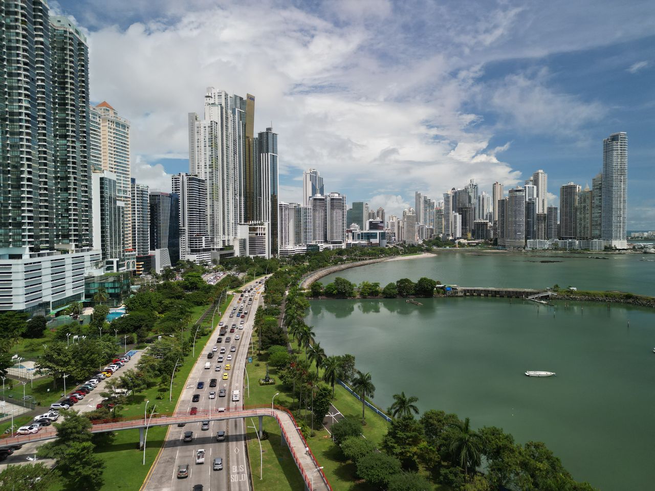 Best Panama eSIM for travel; a panoramic aerial view of  Panama City skyline with tall skyscrapers, a busy highway with cars, green parklands, and a large body of water under a blue sky with white clouds.