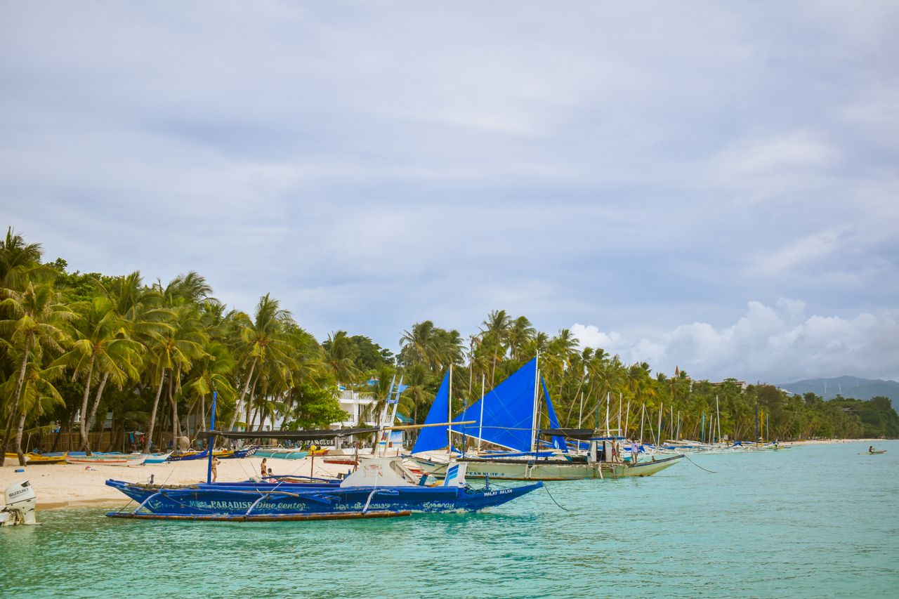 Best eSIM for Philippines; a tropical beach in the Philippines with turquoise water, palm trees, and several traditional blue outrigger boats at anchor.