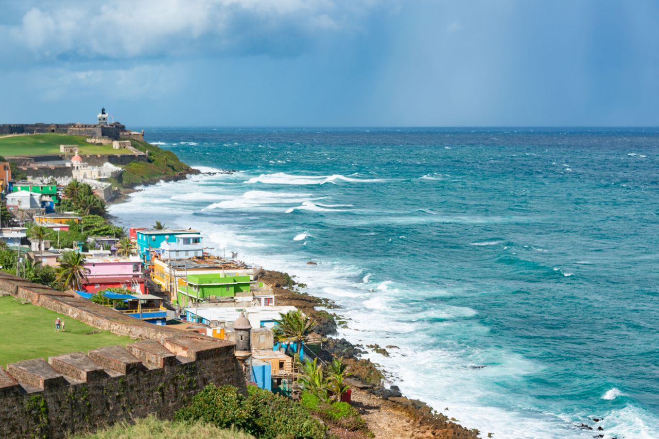 Best Puerto Rico eSIM for travel; coastal landscape in Puerto Rico with turquoise ocean waves crashing against a rocky shore, colorful buildings, green grass, and a cloudy blue sky. 