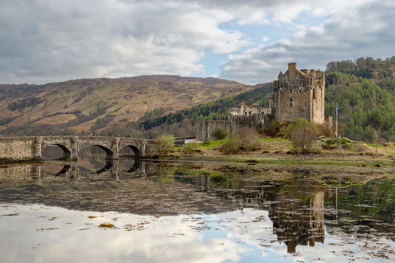 Best eSIM for Scotland; a photograph of a stone castle in Scotland and bridge reflected in calm water, with hills under a cloudy sky.