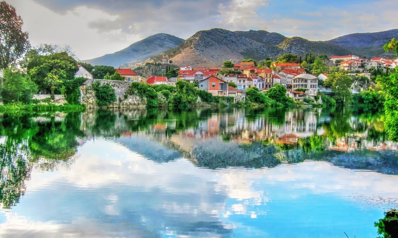 Best eSIM for Serbia; a scenic landscape print showing a small town in Serbia with red-roofed houses along a calm river, with mountains in the background and the entire scene beautifully reflected in the water.
