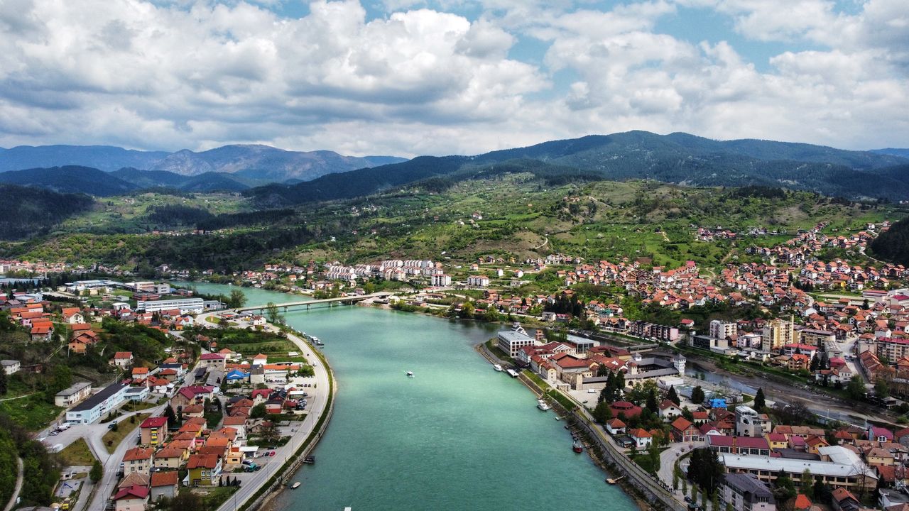 Best Serbia eSIM for travel; aerial view of a town in Serbia situated on a river bend, with green hills, mountains in the background, and a partly cloudy blue sky. 
