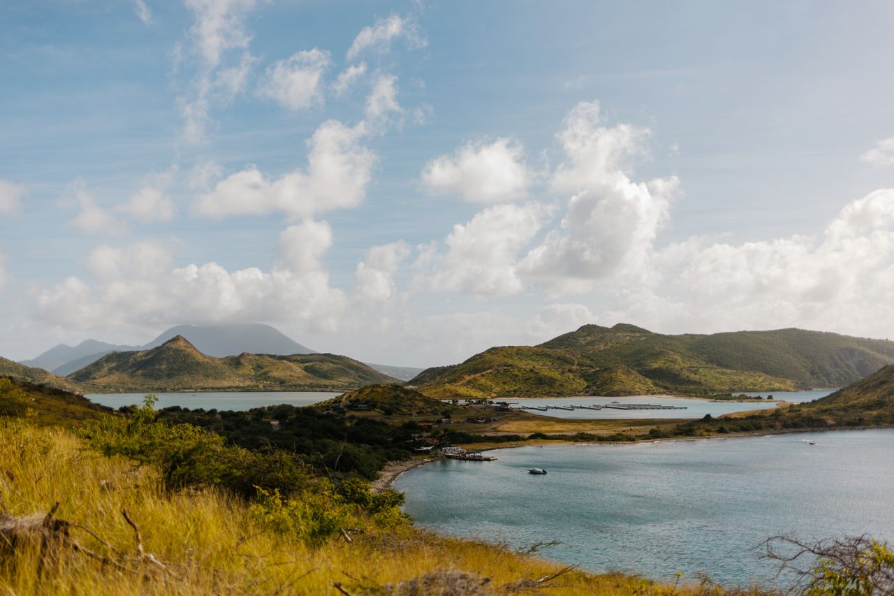 Best Saint Barthelemy eSIM for travel; a scenic landscape in Saint Bathelemy of coastal hills, bays, and water under a blue sky with scattered white clouds. The foreground features dry yellow grasses and green shrubs.