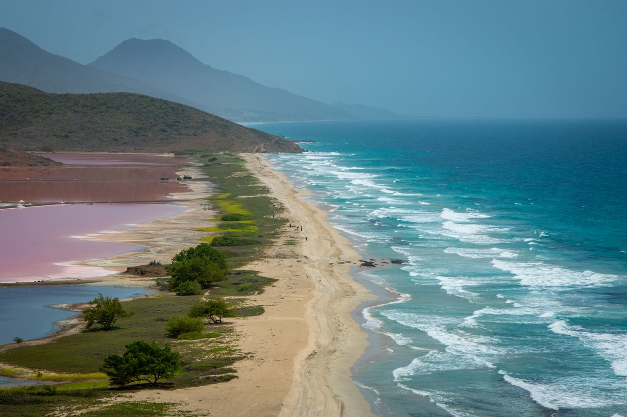 Best eSIM for St. Kitts and Nevis; a coastal landscape with a long sandy beach running between deep blue ocean water and pink salt flats, with hazy mountains in the background.