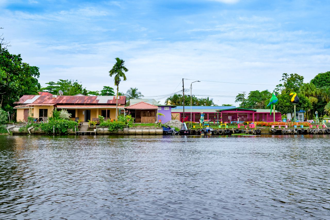 Best Suriname eSIM for travel; A scenic view of a canal in Surname with houses built along the water, surrounded by green trees and a blue sky with light clouds.