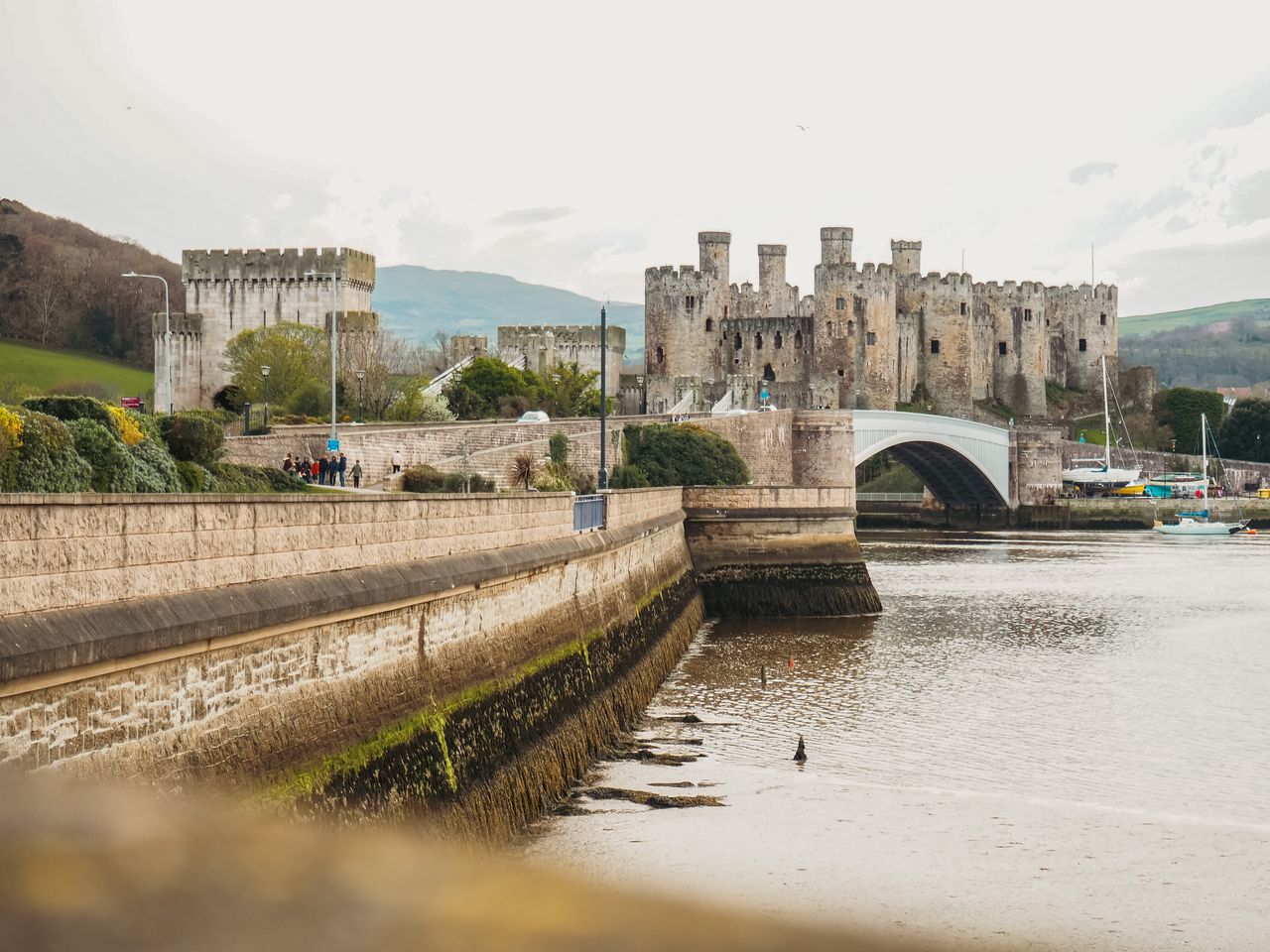 Best eSIM for Wales; a large stone castle in Wales with towers and an arched bridge over calm water, with a stone quay in the foreground and hills in the background.