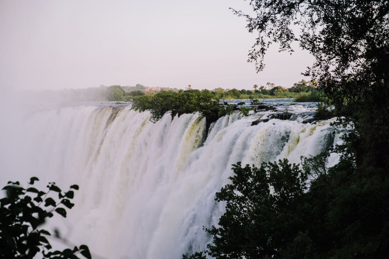Best eSIM for Zambia; a scenic view of a large waterfall cascading over a rocky cliff in Zambia, framed by dark green tree branches and leaves.